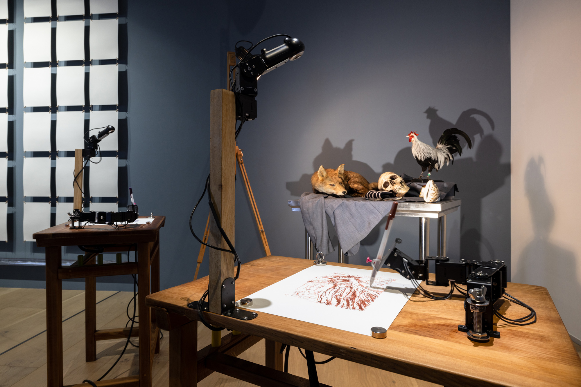 面對狐狸、公雞等標本，三隻機器手臂正在繪製素描。連接機器手臂的攝影鏡頭還會抬頭觀看靜物，再低頭作畫，跟真人的舉動如出一轍。（圖片來源：忠泰美術館提供）